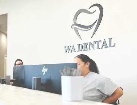Reception area of WA Dental, featuring two receptionists working behind a counter with a prominent wall logo of a stylized tooth above the clinic’s name - Dentist Federal Way WA Reception area of WA Dental, featuring two receptionists working behind a counter with a prominent wall logo of a stylized tooth above the clinic’s name - Dentist Federal Way WA