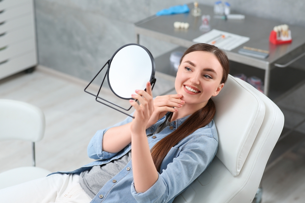 Young woman looking at her new dental implants in mirror indoors - General Dentistry Young woman looking at her new dental implants in mirror indoors - General Dentistry